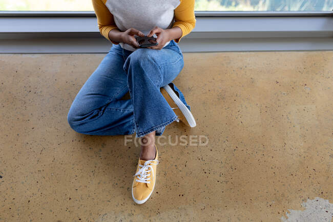 Sección baja de mensajería de texto de mujer de negocios a través de teléfono inteligente mientras está sentado en el suelo en la oficina. negocio independiente creativo, tecnología inalámbrica y oficina. - foto de stock