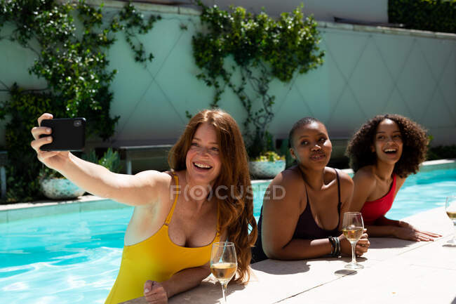 Jovem feliz tomando selfie com amigos femininos multirraciais na piscina no dia ensolarado. amizade, socialização, tecnologia sem fio e tempo de lazer. — Fotografia de Stock