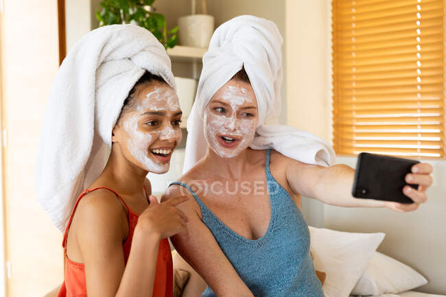 Freundinnen mit Gesichtsmasken und um Haare gewickelten Handtüchern machen zu Hause ein Selfie mit dem Smartphone. häuslicher Lebensstil, Freundschaft, drahtlose Technologie und Hautpflege. — Stockfoto