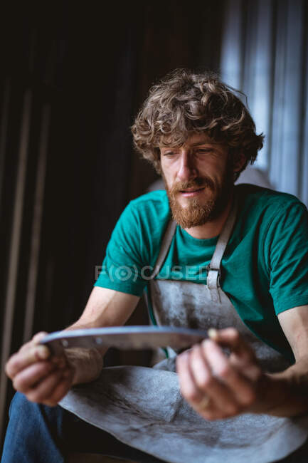 Bearded caucasian blacksmith holding knife shaped metal while sitting in industry. forging, metalwork and manufacturing industry. — Stock Photo