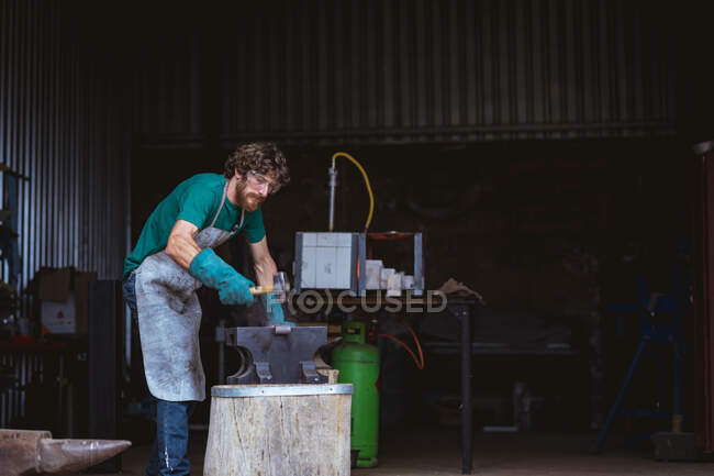 Forgeron caucasien en vêtements de travail protecteurs forgeant avec marteau sur enclume dans l'industrie. forgeage, métallurgie et industrie manufacturière. — Photo de stock
