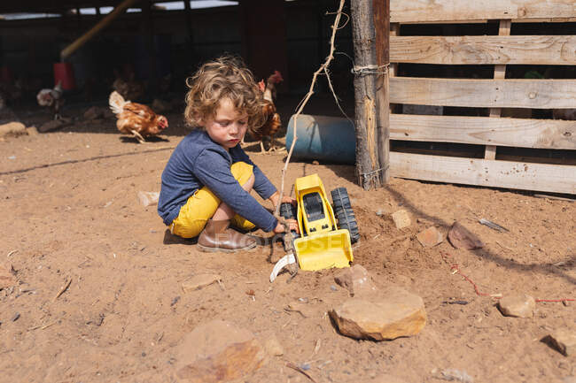 Full length of cute boy crouching while playing with toy bulldozer outside pen. childhood, homesteading and poultry farming. — Stock Photo