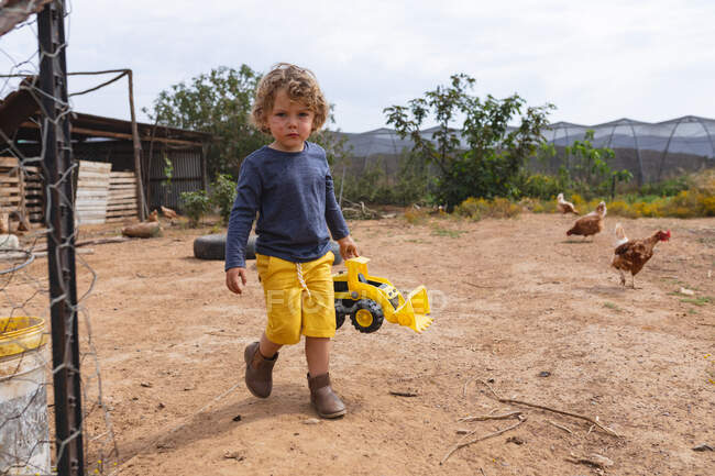 Full length portrait of cute boy holding toy bulldozer while walking outside pen at farm. childhood, homesteading and poultry farming. — Stock Photo