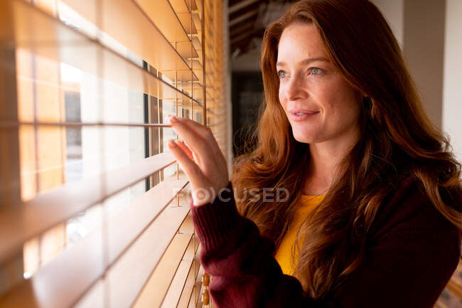 Mujer joven caucásica reflexiva en casuales mirando a través de persianas de la ventana en casa. estilo de vida doméstico y pasar tiempo en casa. - foto de stock
