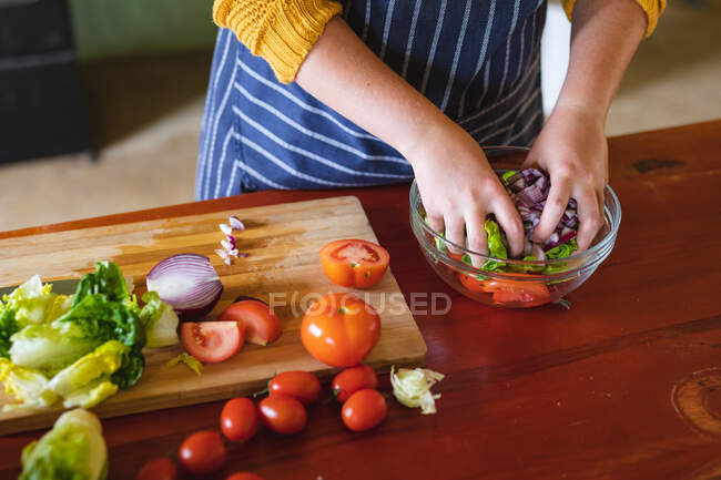 Sección media de la mujer joven mezclando verduras recién picadas en un recipiente de vidrio en el mostrador de la cocina. estilo de vida doméstico y alimentación saludable. - foto de stock
