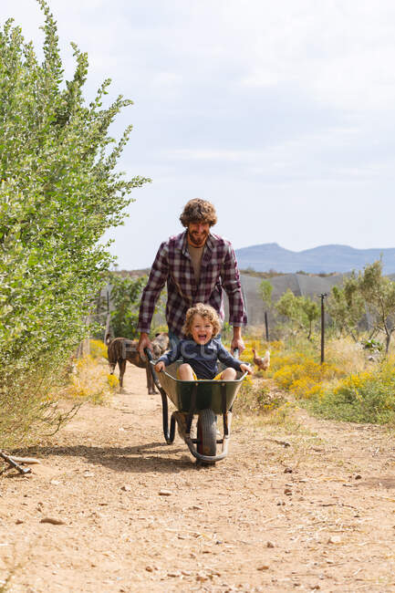 Jovem empurrando garoto alegre e animado sentado no carrinho de mão na passarela na fazenda. família, propriedade e prazer. — Fotografia de Stock