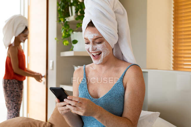 Jovem feliz com máscara facial e toalha embrulhada no cabelo usando smartphone em casa. estilo de vida doméstico, tecnologia sem fio e cuidados com a pele. — Fotografia de Stock