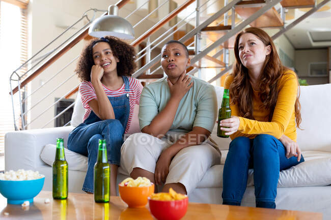 Amigas multirraciais com cerveja e pipocas assistindo TV em casa. amizade, socialização e lazer em casa. — Fotografia de Stock