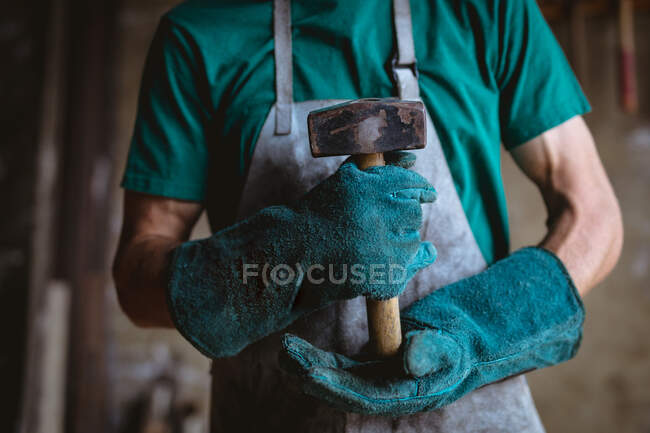 Midsection of blacksmith in protective workwear holding hammer while standing at industry. forging, metalwork and manufacturing industry. — Stock Photo
