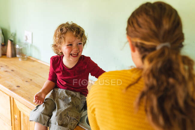 Alegre chico rubio lindo sentado en el mostrador de la cocina mientras se ríe y mira a la madre. estilo de vida familiar y doméstico, infancia. - foto de stock