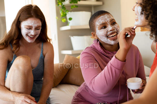 Boas amigas biraciais com máscaras faciais passando o tempo de lazer juntas em casa no fim de semana. amizade, socialização e cuidados com a pele. — Fotografia de Stock