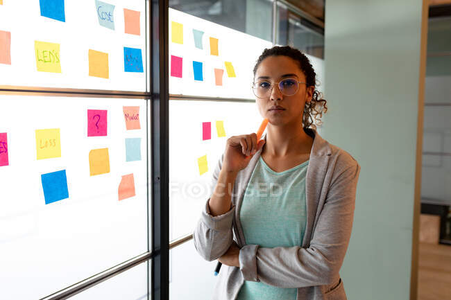 Retrato de empresária birracial confiante em vítimas inteligentes por notas pegajosas na parede no escritório. negócio criativo, escritório moderno e plano de negócios. — Fotografia de Stock