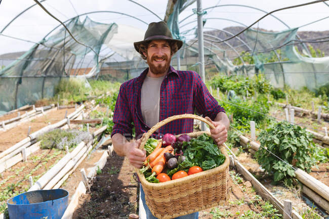 Портрет улыбающегося бородатого мужчины в шляпе со свежесобранными овощами с органической фермы. усадьба и занятие фермерством. — стоковое фото