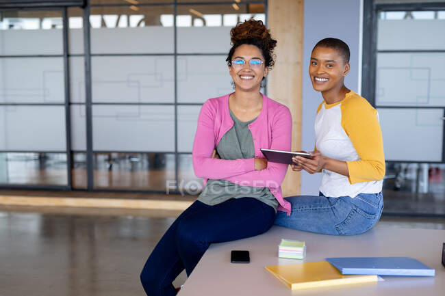 Portrait of smiling multiracial businesswomen in casuals sitting on desk in creative office. creative business, modern office and wireless technology. — Stock Photo