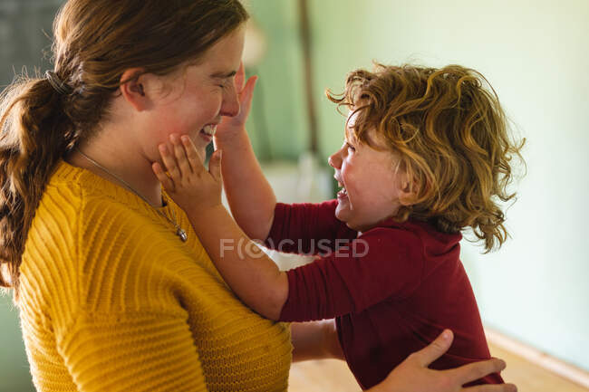 Fröhlicher blonder Junge beim Spielen, während er zu Hause in der Küche die Wange der Mutter berührt. Kindheit, Familie und häuslicher Lebensstil. — Stockfoto