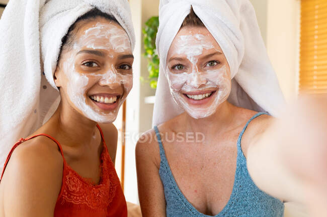 Cheerful female friends with facial masks and towels wrapped on hair taking selfie at home. domestic lifestyle, friendship and skincare. — Stock Photo