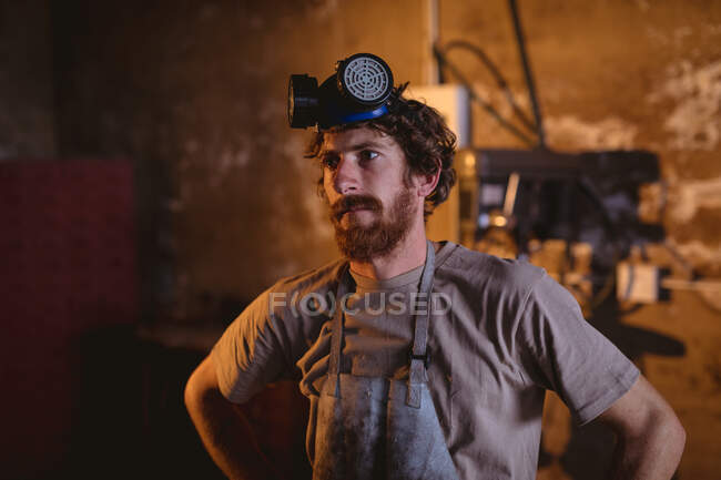 Thoughtful bearded caucasian blacksmith with gas mask looking away while standing in industry. forging, metalwork and manufacturing industry. — Stock Photo