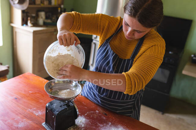 Junge Frau gießt Mehl aus Behälter auf Gewichtswaage am Tisch in der Küche. Lebensstil und gesunde Ernährung. — Stockfoto