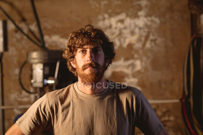 Portrait of bearded caucasian blacksmith in t-shirt standing in metal industry. forging, metalwork and manufacturing industry. — Stock Photo