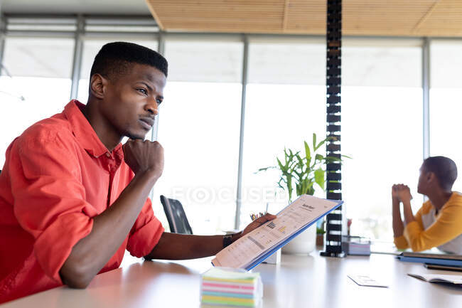 Pensativo empresário afro-americano casual com a mão no queixo segurando documento no escritório. negócios criativos e escritório moderno. — Fotografia de Stock