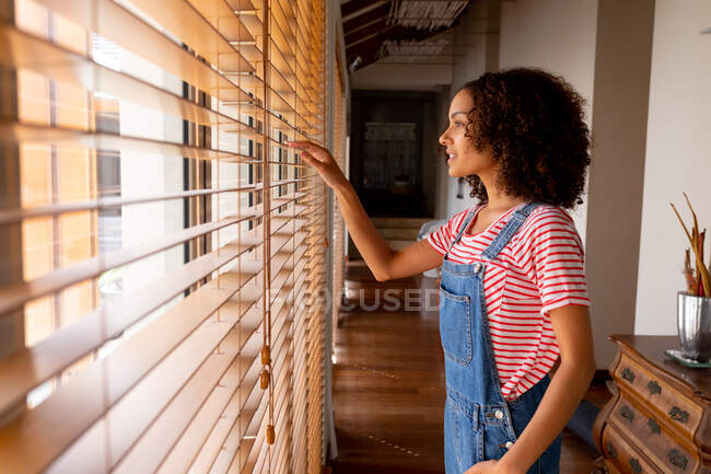 Thoughtful biracial woman in bib overalls looking through window blinds at home. domestic lifestyle and spending time at home. — Stock Photo