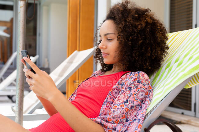 Biracial young woman in swimsuit using smart phone while relaxing on deck chair at poolside. lifestyle, leisure time and weekend at poolside. — Stock Photo