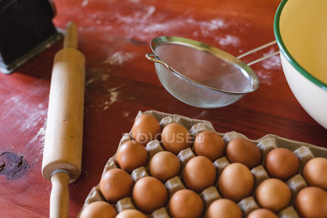 Vista de ángulo alto de los huevos marrones en el cartón por rodillo y colador en la mesa de madera en casa. alimentación orgánica y saludable. - foto de stock