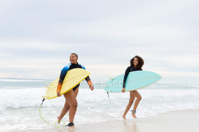 Des amies souriantes multiraciales avec des planches de surf courant au bord de la mer contre le ciel pendant le week-end. amitié, surf et loisirs. — Photo de stock