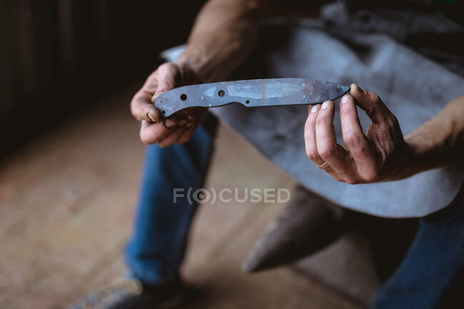 Sección baja de herrero sosteniendo cuchillo en forma de metal mientras está sentado en la industria. forja, metalurgia e industria manufacturera. - foto de stock