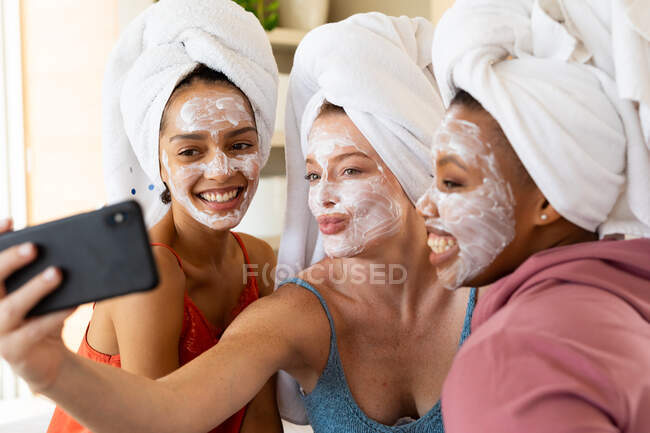 Mujeres jóvenes felices con máscaras faciales y toallas envueltas en el pelo tomando selfie en casa. amistad, cuidado de la piel, tecnología inalámbrica y tiempo libre. - foto de stock