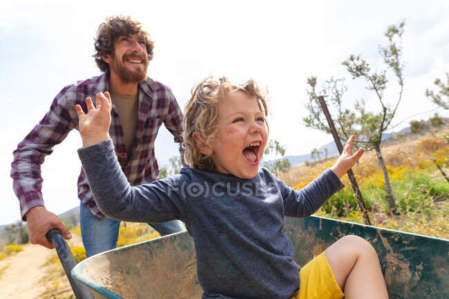 Feliz joven empujando excitado hijo gritando mientras está sentado en carretilla en la granja. familia, hacienda y disfrute. - foto de stock