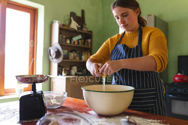 Jovem mulher quebrando casca de ovo na tigela enquanto prepara a massa na mesa na cozinha. estilo de vida doméstico e alimentação saudável. — Fotografia de Stock
