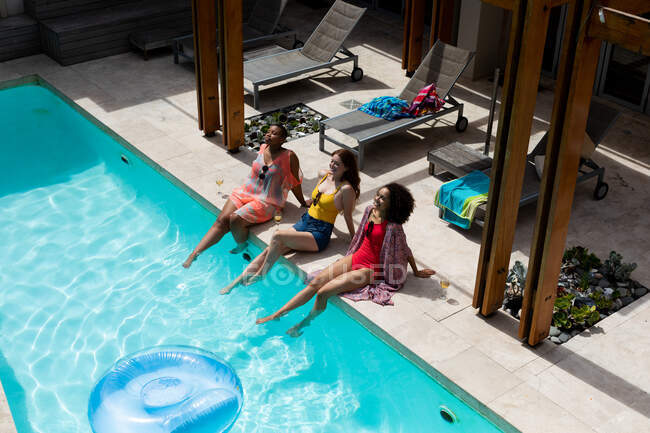 Счастливые многорасовые подруги загорают в бассейне в выходные в солнечный день. дружба, общение и досуг. — стоковое фото