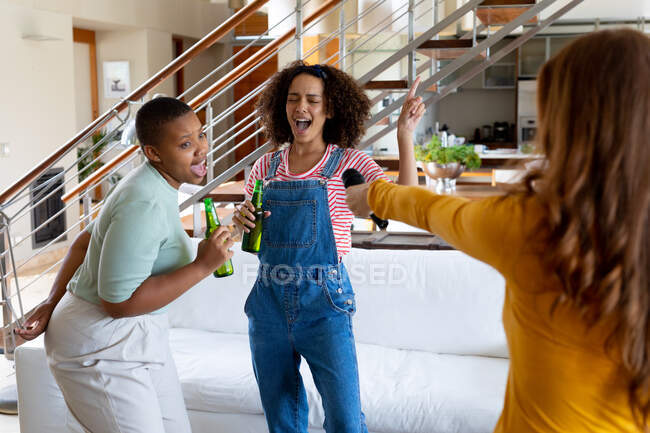 Cheerful multiracial female friends singing and having fun while holding beer bottles at home. friendship, socialising and leisure time at home. — Stock Photo