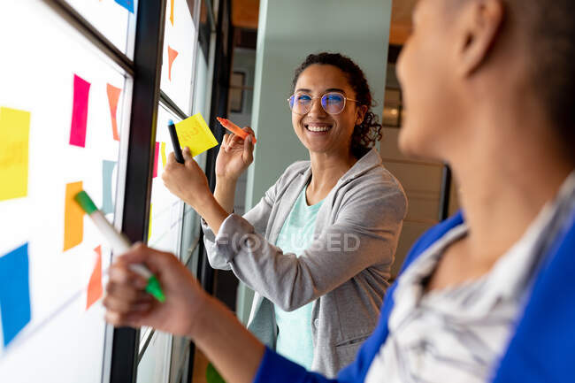 Mujer de negocios birracial sonriente con colega que planea la estrategia de negocios sobre notas adhesivas en la oficina. negocio creativo, oficina moderna y plan de negocio. - foto de stock