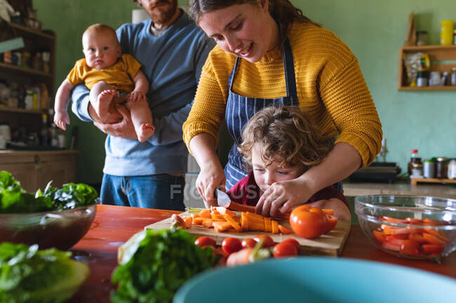 Frau und Sohn schneiden frische Möhren, während der Mann das Baby zu Hause in der Küche trägt. Familie, gesunde Ernährung und häuslicher Lebensstil. — Stockfoto