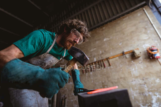 Forgeron caucasien barbu en vêtements de travail protecteurs forgeant avec marteau sur enclume dans l'industrie. forgeage, métallurgie et industrie manufacturière. — Photo de stock
