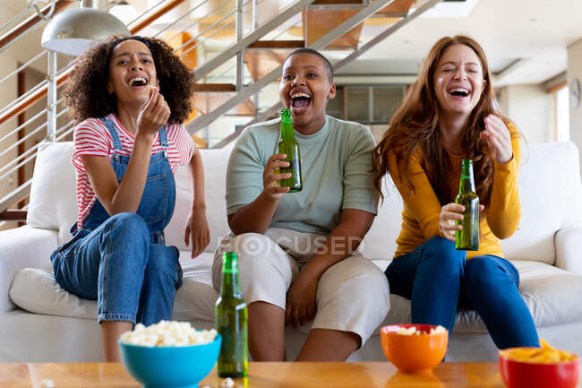 Vielrassige Freundinnen mit Bier und Popcorn lachen, während sie zu Hause fernsehen. Freundschaft, Geselligkeit und Freizeit zu Hause. — Stockfoto