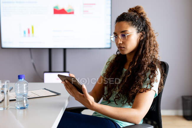 Empresaria birracial de confianza en casuales que utilizan tableta digital mientras está sentado en la oficina creativa. negocios creativos, oficina moderna y tecnología inalámbrica. - foto de stock