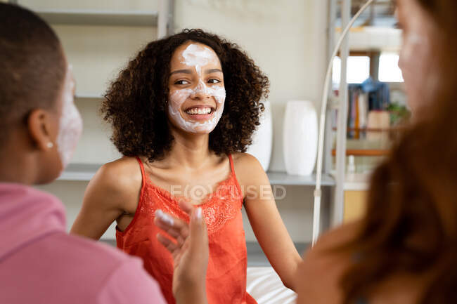 Feliz birracial amigos do sexo feminino com máscaras faciais passar o tempo de lazer em casa durante o fim de semana. amizade, socialização e cuidados com a pele. — Fotografia de Stock