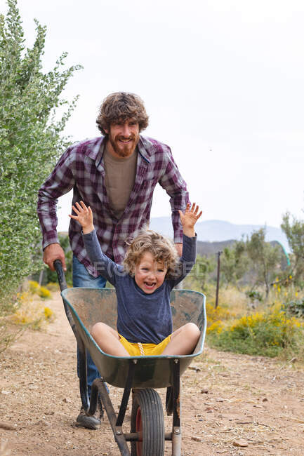 Giovane uomo spingendo figlio allegro ed eccitato seduto con le mani alzate in carriola sulla passerella. famiglia, fattoria e divertimento. — Foto stock