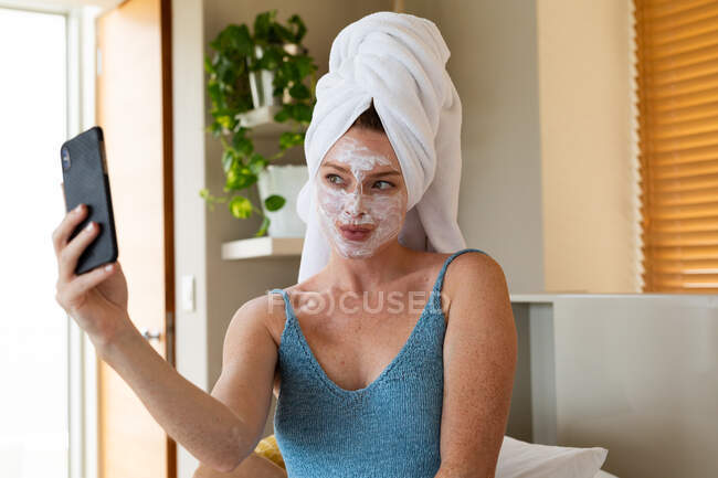 Mujer joven con máscara facial y toalla envuelta en el pelo tomando selfie a través de teléfono inteligente en casa. estilo de vida doméstico, tecnología inalámbrica y cuidado de la piel. - foto de stock