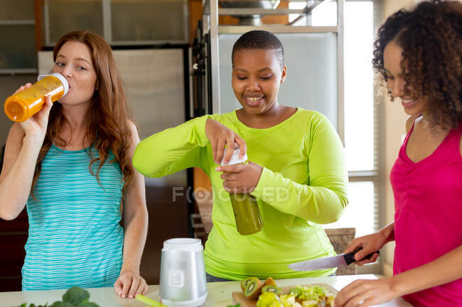 Mulheres multirraciais bebendo suco enquanto sorrindo amigo feminino cortando legumes na ilha da cozinha. amizade, socialização e festa em casa. — Fotografia de Stock