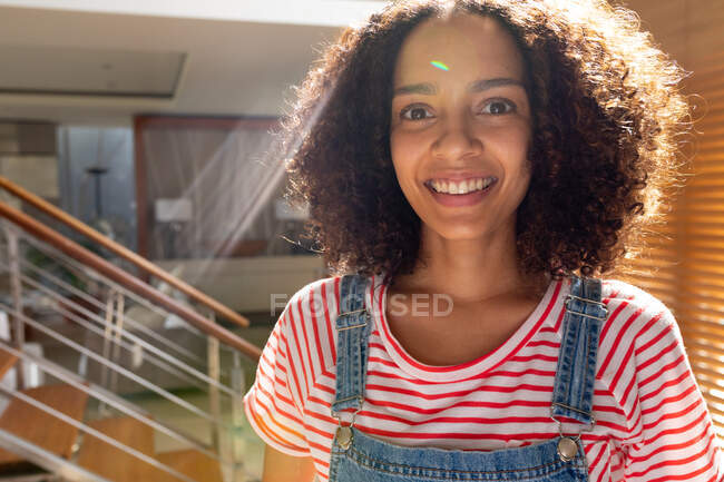Portrait of happy biracial woman in bib overalls at home during sunny day. domestic lifestyle and leisure. — Stock Photo