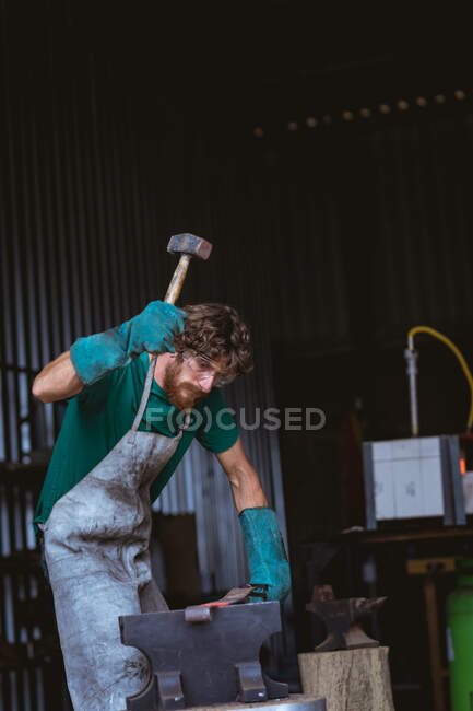 Caucasian bearded blacksmith in protective workwear forging with hammer on anvil in industry. forging, metalwork and manufacturing industry. — Stock Photo