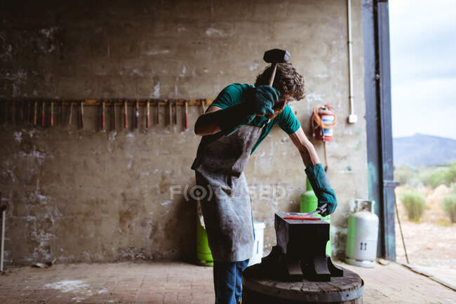 Forgeron caucasien en gants de protection forgeant avec marteau sur enclume dans l'industrie manufacturière. forgeage, métallurgie et industrie manufacturière. — Photo de stock