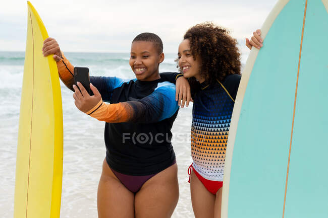 Felices amigas multirraciales con tablas de surf tomando selfie a través de smartphone en la playa. amistad, surf y tiempo libre. - foto de stock