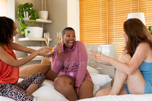 Felices amigas multirraciales disfrutando del champán mientras se sientan en la cama durante la fiesta de la casa. amistad, socialización y tiempo libre. - foto de stock
