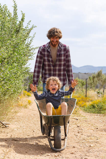 Happy father pushing cheerful son sitting in wheelbarrow on walkway at farm during sunny day. family, homesteading and enjoyment. — Stock Photo