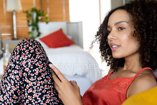 Thoughtful biracial young woman in casuals looking away while holding smart phone at home. domestic lifestyle and leisure. — Stock Photo